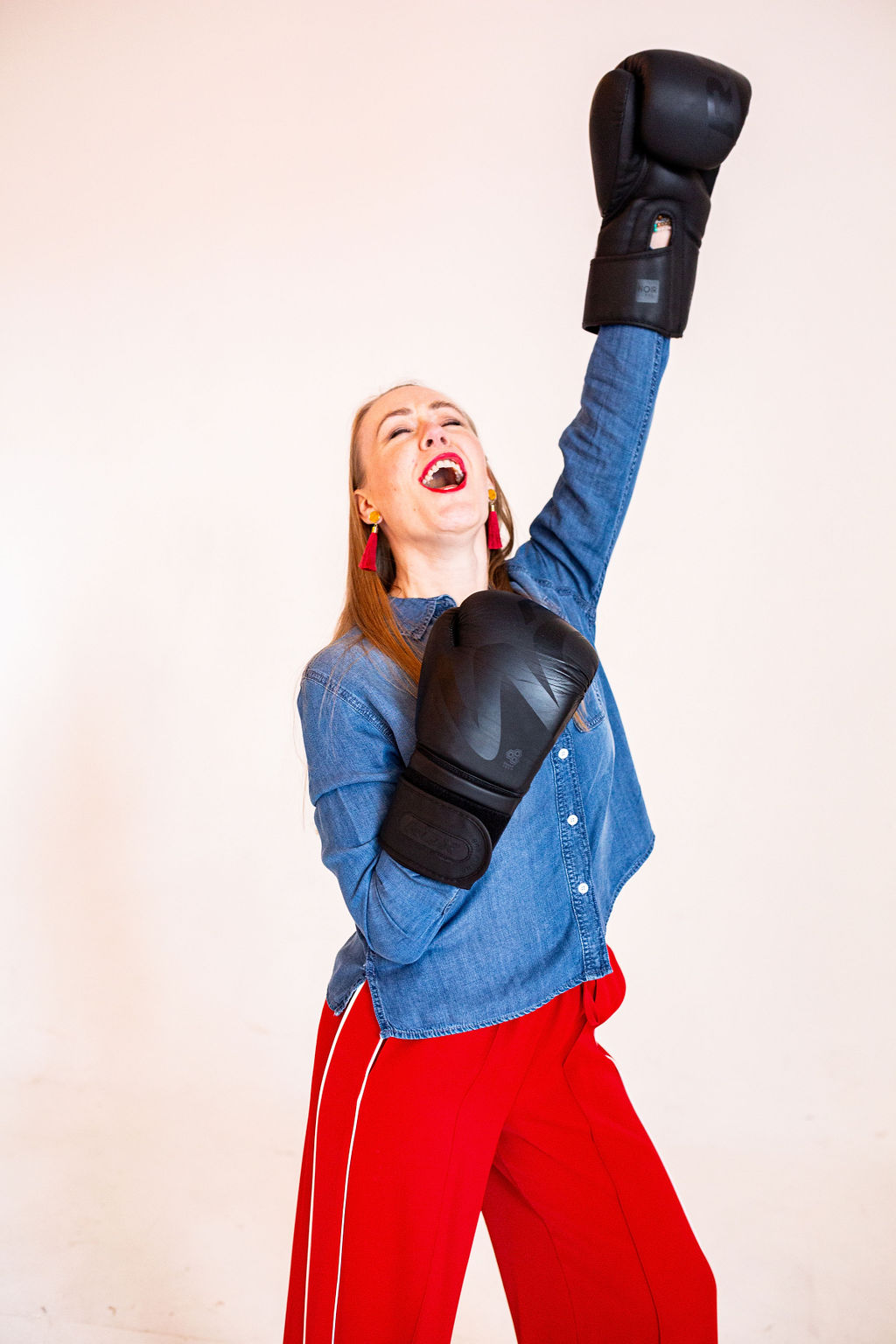 Lindsey Hall throws her head back in joyful triumph, wearing boxing gloves and bright red trousers, mid-laugh with one arm raised in victory. A playful, powerful moment that captures the strength and freedom found on the other side of emotional healing.