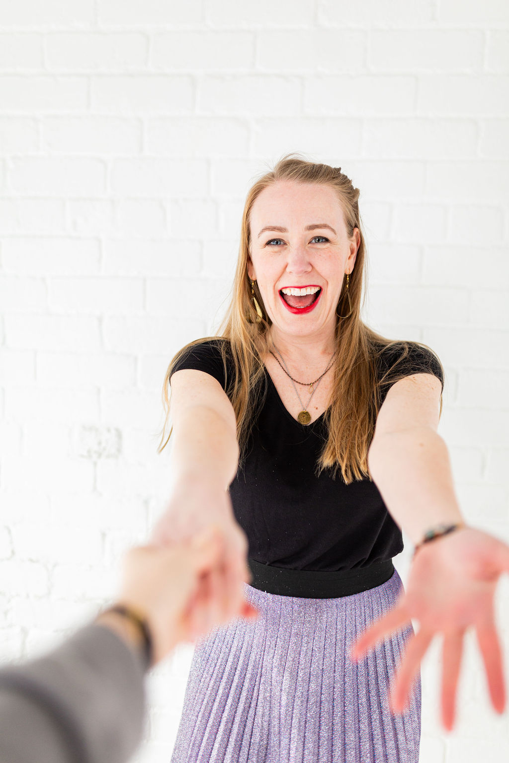 Lindsey Hall, wearing a sparkly purple skirt, smiling brightly with arms outstretched toward the camera, reaching for someone’s hand. A joyful, welcoming moment that radiates warmth, connection, and the spirit of walking this journey together.