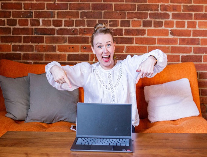 Lindsey wearing white flowy shirt on orange couch, with excited look on her face, pointing down at laptop